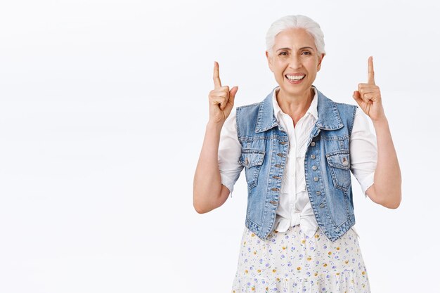 Señora mayor caucásica feliz y despreocupada con cabello gris, sonrisa encantadora y alegre, riendo mientras señala con el dedo hacia arriba, recomendar un producto o promoción, anunciar algo, usar un chaleco de mezclilla con estilo moderno