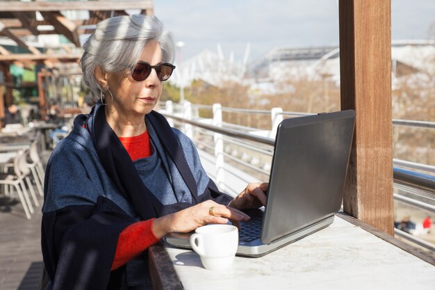 Señora mayor seria que trabaja en la computadora en café al aire libre