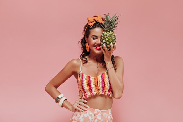 Señora de moda optimista con pañuelo naranja y labios rojos en ropa de verano de moda posando con piña y ojos cerrados.