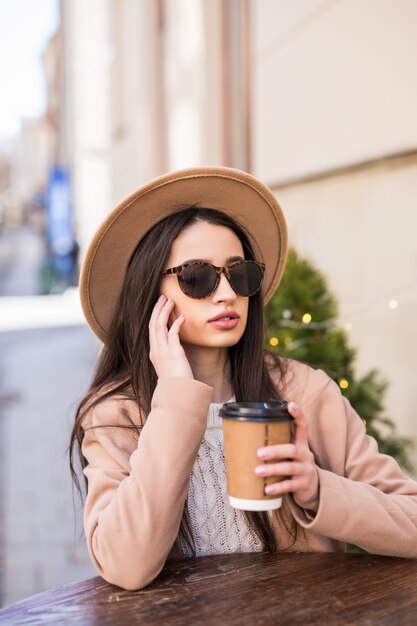 Señora modelo de moda está sentada en la mesa en los vestidos de café en ropa casual gafas de sol oscuras con taza de café