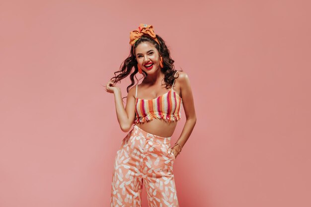 Señora moderna alegre con peinado ondulado y diadema naranja en la parte superior brillante a rayas y pantalones elegantes posando sobre fondo rosa