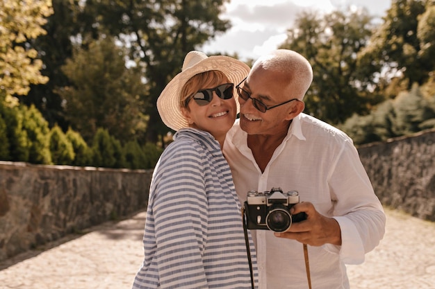 Foto gratuita señora moderna con cabello rubio en sombrero gafas de sol y traje azul a rayas sonriendo con anciano con bigote en camisa blanca con cámara en el parque