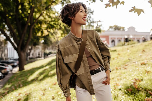 Señora moderna en chaqueta de mezclilla con bolso mirando hacia fuera. Mujer de pelo ondulado con labios rojos en pantalón blanco sonriendo al aire libre.