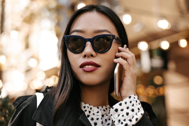 Señora morena bronceada en gafas de sol habla por teléfono Encantadora mujer asiática en traje blanco y negro posa afuera