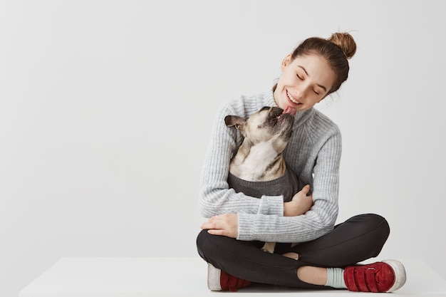 Señora morena contenta en ropa casual sentado en la mesa con perro en las manos. Diseñador de inicio femenino que abraza el perro pedigrí mientras se lame la barbilla. Concepto de alegría, espacio de copia