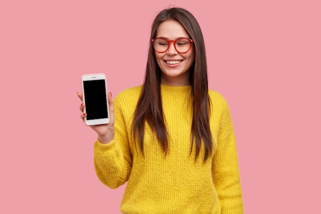 Señora morena optimista muestra la pantalla del teléfono inteligente, se regocija comprando un nuevo dispositivo, usa gafas y un suéter amarillo