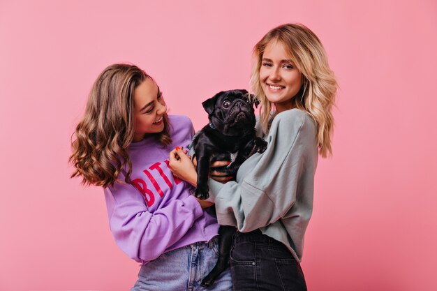 Señora morena rizada en ropa de moda mirando cachorro de bulldog y riendo. Espectaculares chicas caucásicas que pasan tiempo libre con su perro.