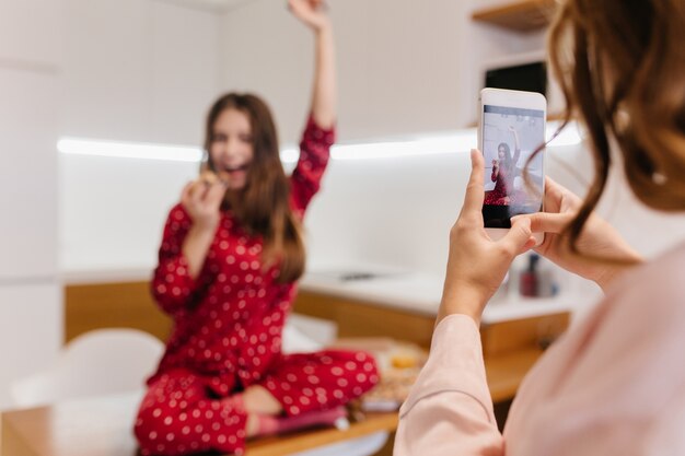 Señora morena sosteniendo el teléfono inteligente mientras toma la foto de su hermana riendo. Mujer joven adorable en ropa de dormir roja sentada con las piernas dobladas y agitando la mano.