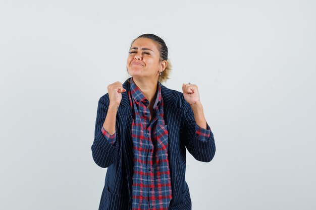 Señora mostrando gesto de ganador en camisa, chaqueta y mirando feliz, vista frontal.