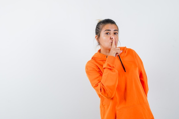 Señora mostrando gesto de silencio en sudadera con capucha naranja y mirando sensible