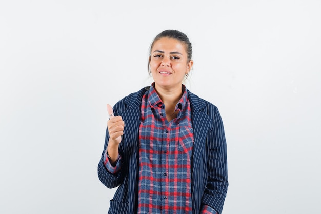 Señora mostrando el pulgar hacia arriba en camisa, chaqueta y mirando confiado, vista frontal.