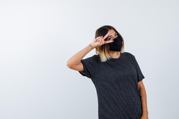 Foto gratuita señora mostrando el signo de la victoria en el ojo con vestido negro, máscara médica y mirando feliz, vista frontal.