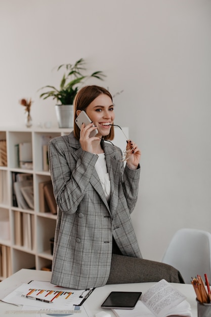 Señora de negocios alegre sonríe mientras se comunica por teléfono. Mujer en chaqueta a cuadros gris posando en oficina blanca.