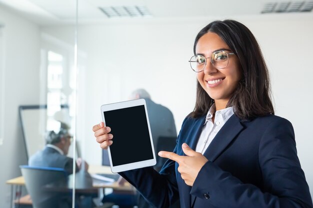Señora de negocios confiada feliz que sostiene la tableta, mostrando y señalando el dedo en la pantalla en blanco, mirando a la cámara y sonriendo. Copie el espacio. Concepto de comunicación y publicidad