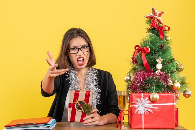 Señora de negocios confundida en traje con gafas sosteniendo su regalo pidiendo algo y sentado en una mesa con un árbol de Navidad en la oficina