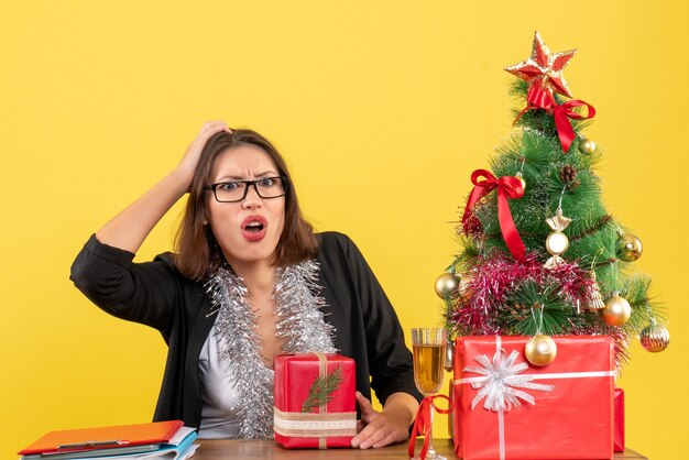 Señora de negocios confundida en traje con gafas sosteniendo su regalo y sentada en una mesa con un árbol de Navidad en la oficina