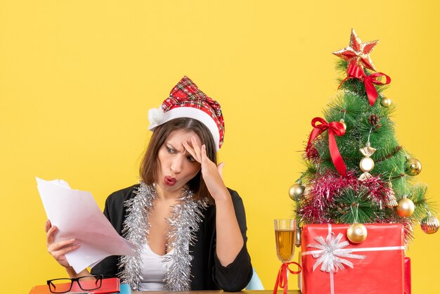 Señora de negocios confundida en traje con sombrero de santa claus y adornos de año nuevo sosteniendo documentos y sentado en una mesa con un árbol de Navidad en la oficina