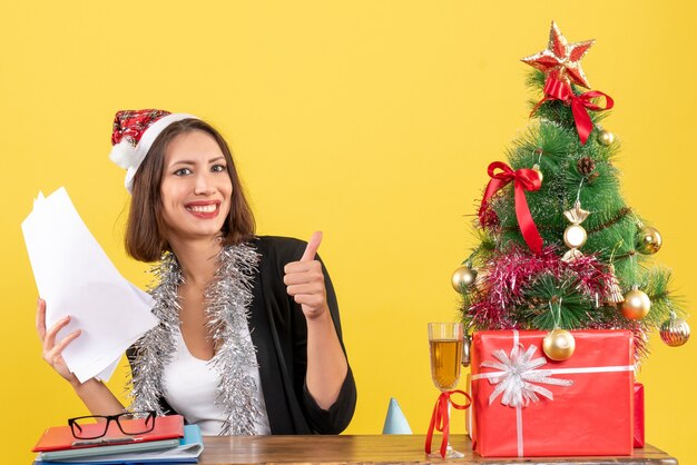 Señora de negocios emocional satisfecha en traje con sombrero de santa claus y adornos de año nuevo sosteniendo documentos haciendo un gesto aceptable y sentado en una mesa con un árbol de Navidad en la oficina