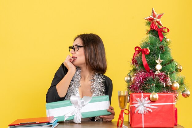 Señora de negocios emocional en traje con gafas apuntando su regalo y sentado en una mesa con un árbol de Navidad en la oficina