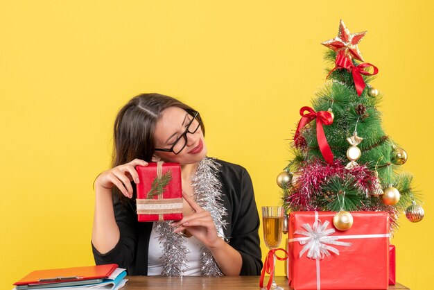 Señora de negocios de ensueño en traje con gafas sosteniendo su regalo y sentada en una mesa con un árbol de Navidad en la oficina