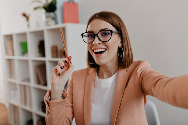 Señora de negocios con gafas y elegante traje ligero hace selfie, sosteniendo un lápiz naranja en sus manos.