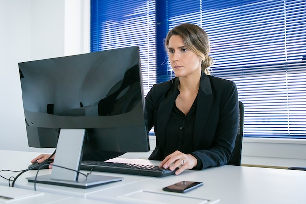 Señora de negocios joven enfocada que trabaja en su oficina, usando la computadora en el lugar de trabajo, mirando la pantalla. Tiro medio. Concepto de líder empresarial o comunicación digital