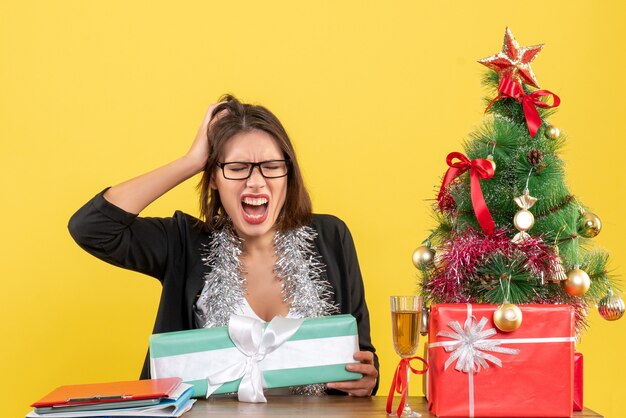 Señora de negocios nerviosa emocional en traje con gafas sosteniendo su regalo y sentada en una mesa con un árbol de Navidad en la oficina