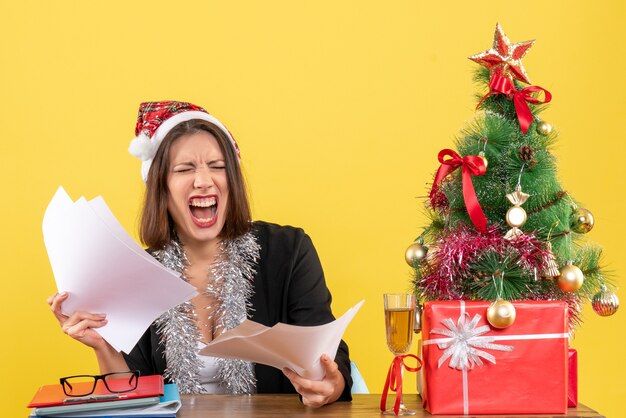 Señora de negocios nerviosa emocional en traje con sombrero de santa claus y decoraciones de año nuevo sosteniendo documentos y sentado en una mesa con un árbol de Navidad en la oficina