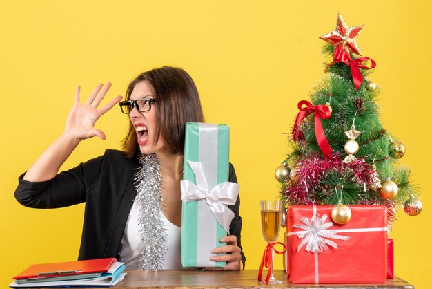 Señora de negocios nerviosa enojada en traje con gafas mostrando su regalo y sentado en una mesa con un árbol de Navidad en la oficina