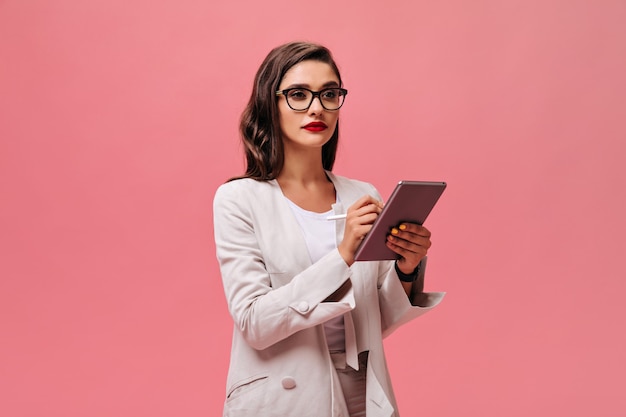 Señora de negocios serio con labios rojos brillantes en traje elegante beige y anteojos sostiene tableta sobre fondo rosa aislado.