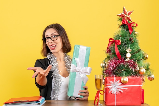 Señora de negocios sonriente en traje con gafas mostrando su regalo pidiendo algo y sentado en una mesa con un árbol de Navidad en la oficina