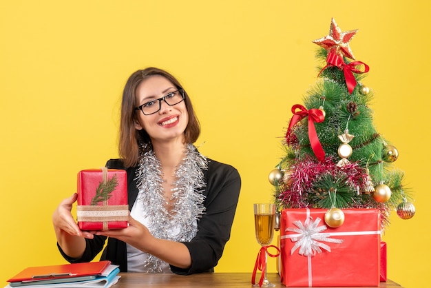 Señora de negocios sonriente en traje con gafas mostrando su regalo y sentado en una mesa con un árbol de Navidad en la oficina