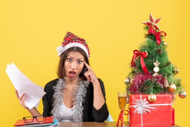 Señora de negocios sorprendida en traje con sombrero de santa claus y adornos de año nuevo sosteniendo documentos y sentado en una mesa con un árbol de Navidad en la oficina