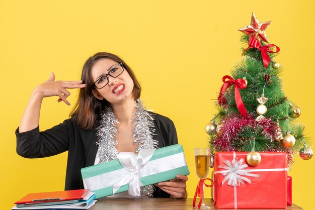 Señora de negocios en traje con gafas mostrando su regalo confundida por algo y sentada en una mesa con un árbol de Navidad en la oficina