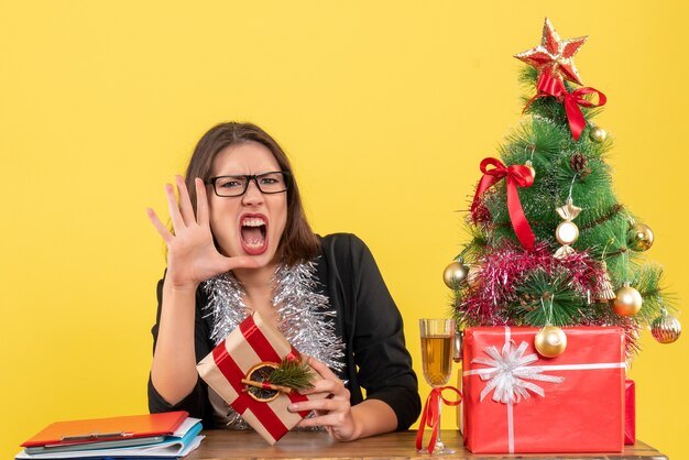 Señora de negocios en traje con gafas sosteniendo su regalo gritando a alguien y sentada en una mesa con un árbol de Navidad en la oficina