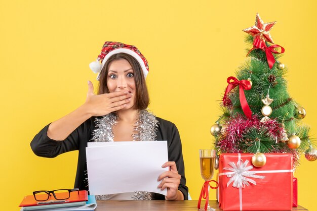 Señora de negocios en traje con sombrero de santa claus y adornos de año nuevo mirando algo sorprendentemente y sentado en una mesa con un árbol de Navidad en la oficina