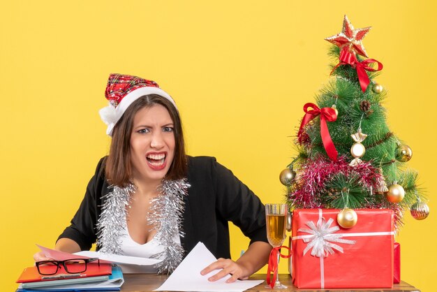 Señora de negocios en traje con sombrero de santa claus y adornos de año nuevo que se siente enojada y sentada en una mesa con un árbol de Navidad en la oficina