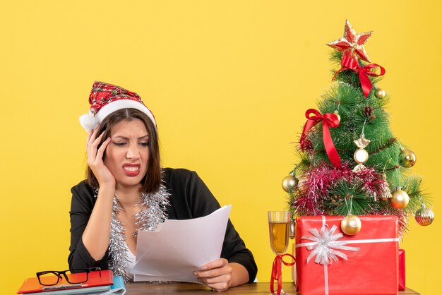 Señora de negocios en traje con sombrero de santa claus y adornos de año nuevo sintiéndose agotada y sentada en una mesa con un árbol de Navidad en la oficina