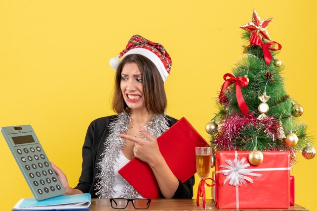 Señora de negocios en traje con sombrero de santa claus y decoraciones de año nuevo sintiéndose confundida mirando calculadora y sentada en una mesa con un árbol de Navidad en la oficina