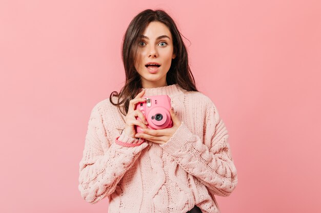 Señora de ojos grises con expresión de asombro hace una foto en una mini cámara rosa. Mujer con cabello lacio oscuro en suéter de punto posando sobre un fondo aislado.