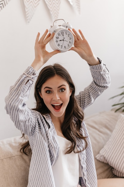 Foto gratuita señora de ojos verdes en pijama posando felizmente en el apartamento y sosteniendo el despertador