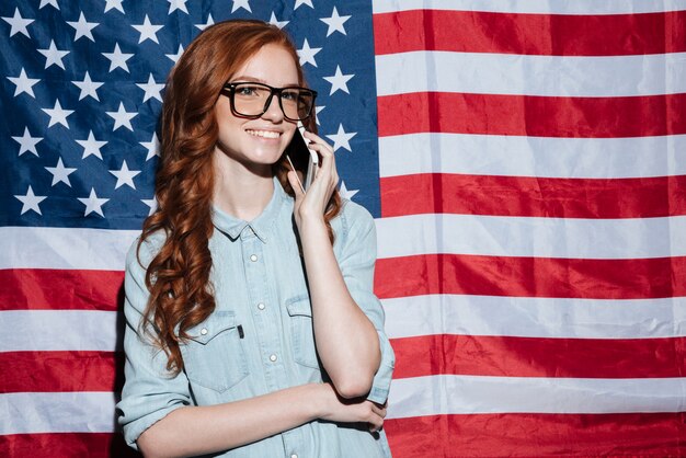 Señora pelirroja alegre de pie sobre la bandera de Estados Unidos hablando por teléfono.