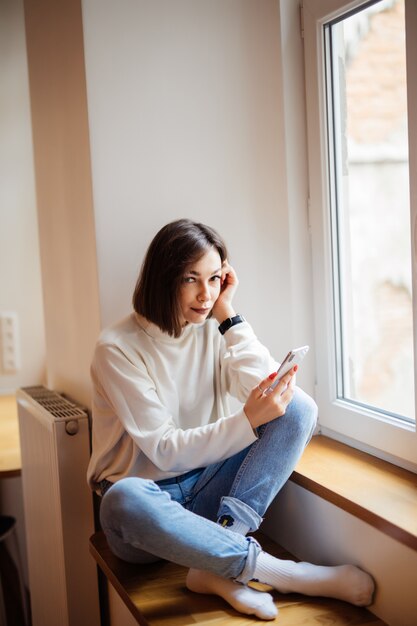 Señora de pelo corto en blue jeans sentado en el alféizar de la ventana y escribir un mensaje en el teléfono inteligente en casa