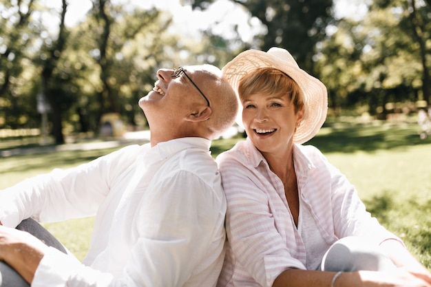 Señora de pelo corto de moda con sombrero ligero y blusa a rayas sonriendo y sentado en la hierba con el anciano con gafas y camisa blanca al aire libre.