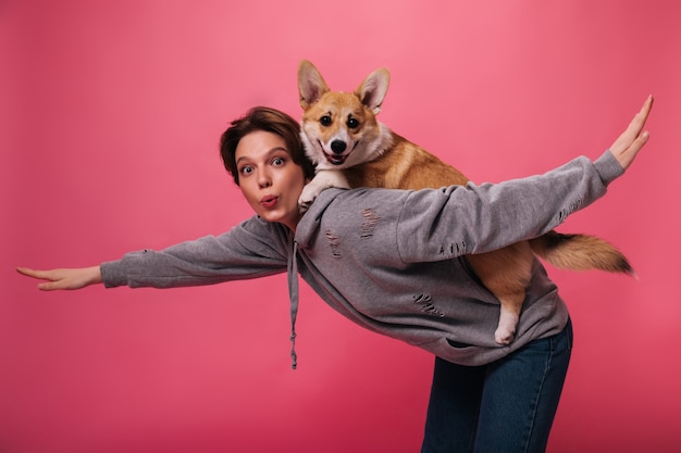 Señora de pelo corto en sudadera con capucha sostiene y juega con perro. Cool mujer en sudadera gris y jeans posa con corgi sobre fondo rosa aislado