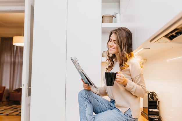 Señora de pelo oscuro alegre pasar la mañana en casa, leyendo el periódico con una sonrisa