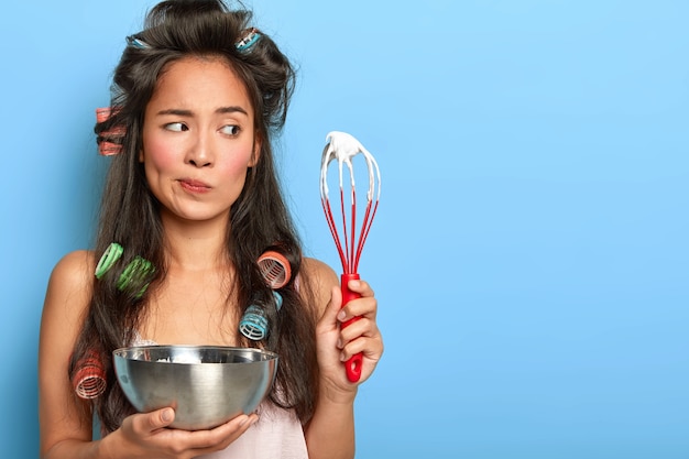 Foto gratuita señora pensativa de cabello oscuro ocupada cocinando postre, sostiene un batidor y un tazón, mezcla huevo blanco para hacer crema, usa rizadores de pelo, ropa de dormir, tiene expresión facial indecisa, posa sobre una pared azul