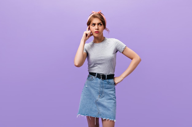 Señora pensativa en poses de falda de mezclilla sobre fondo aislado. Mujer joven con estilo en diadema rosa y camisa ligera mirando a otro lado.