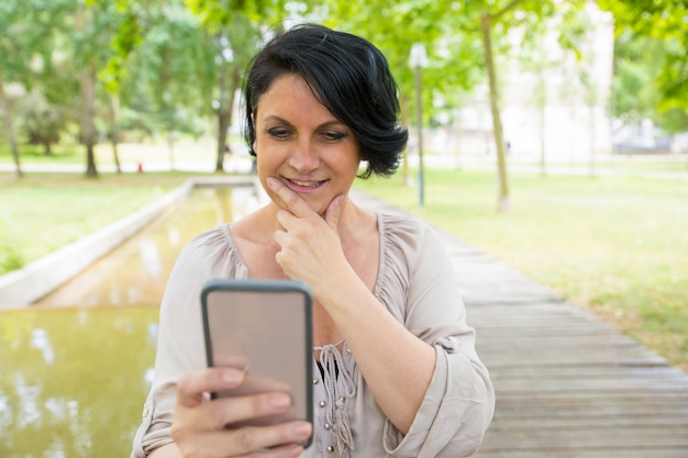 Señora pensativa sonriente tomando fotos en el teléfono inteligente