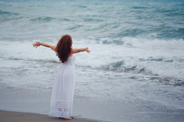 Señora de pie mirando sewith manos abiertas en vestido largo y blanco en la playa durante el día.